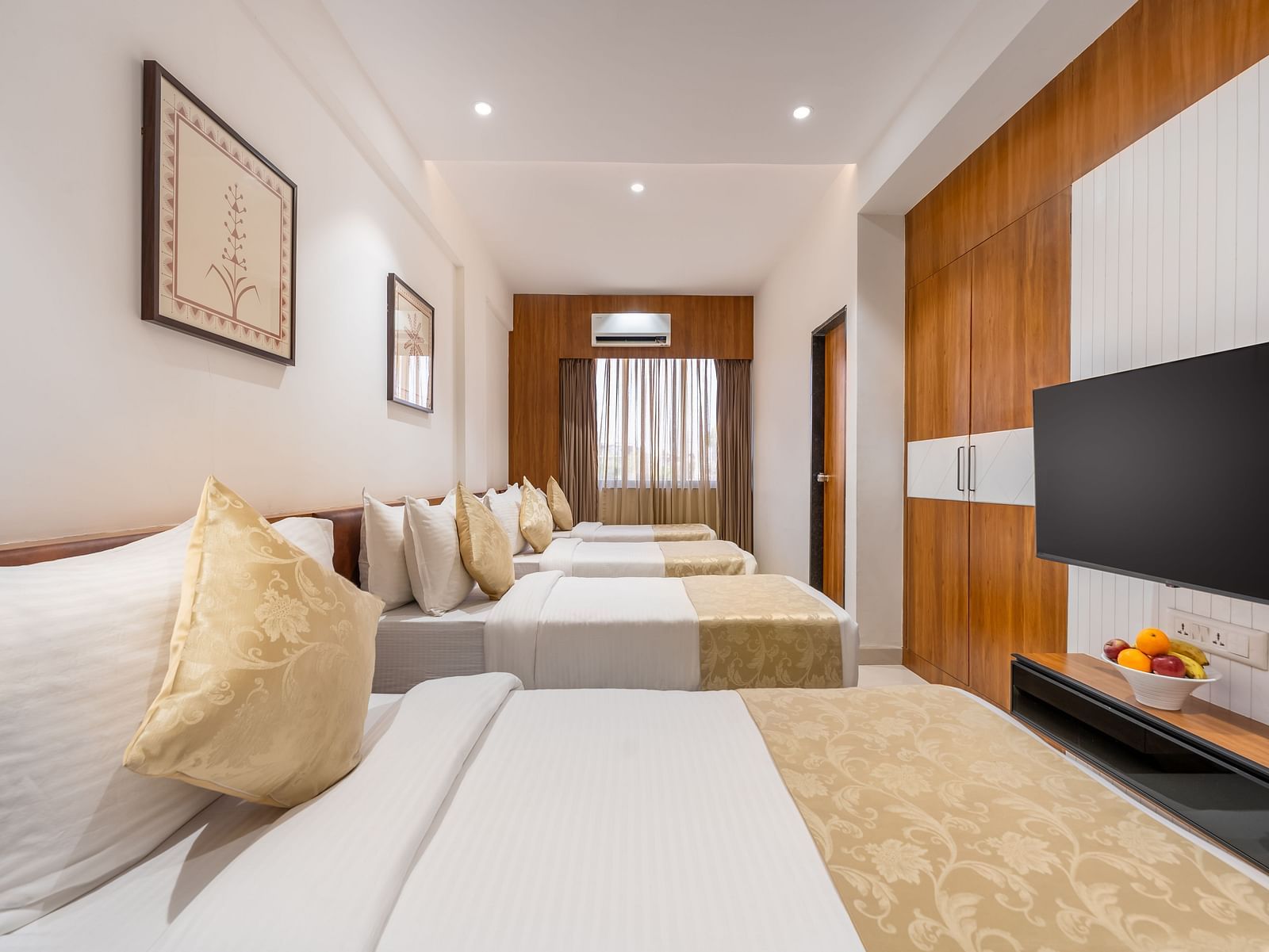 A hotel room contains four neatly made single beds, a mounted television, and simple framed artwork on the walls - Lords Eco Inn Ambaji.