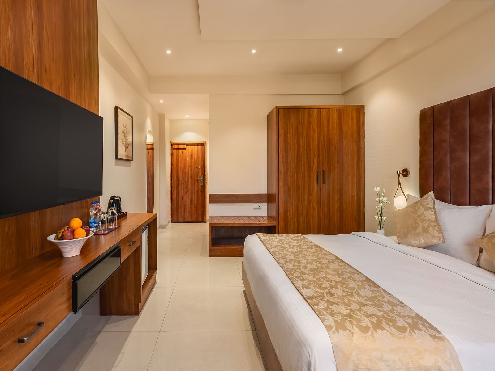 A well-appointed hotel room features a comfortable king-size bed, a mounted television above a desk with fruit, and a wooden wardrobe - Lords Eco Inn Ambaji.