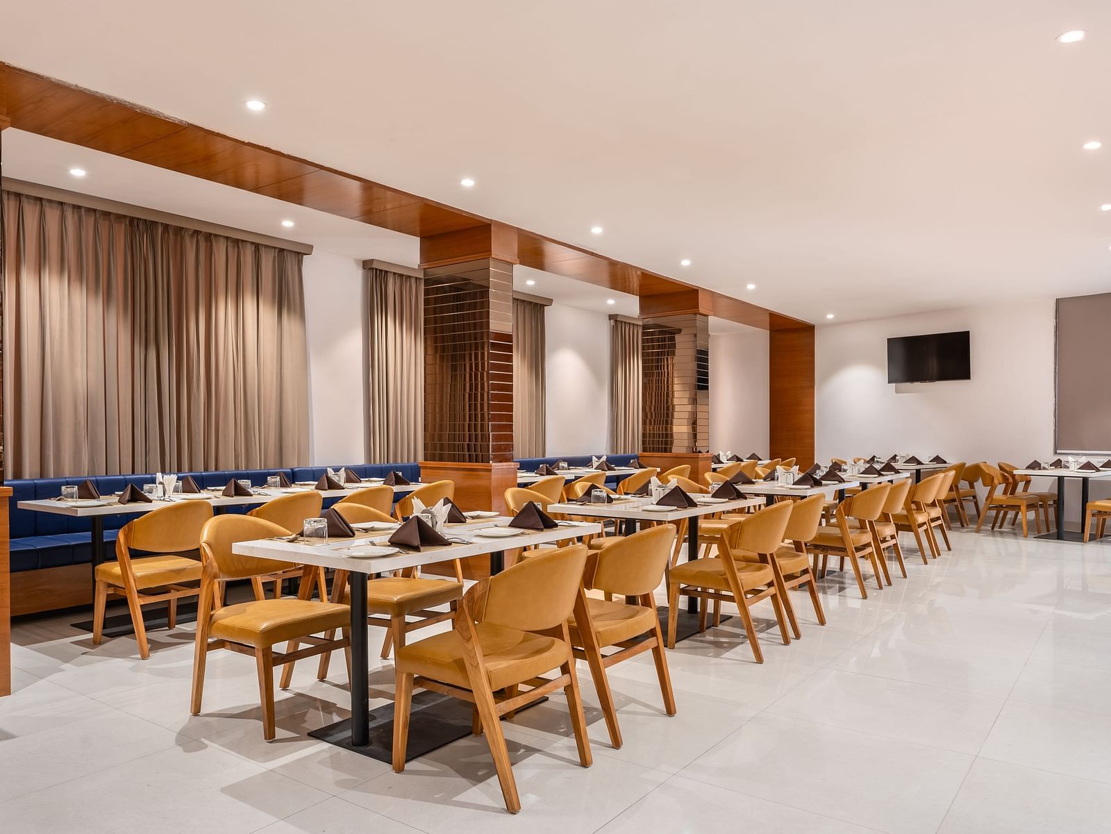 A brightly lit dining area features numerous set tables with chairs and booth seating along the wall - Lords Eco Inn Ambaji.