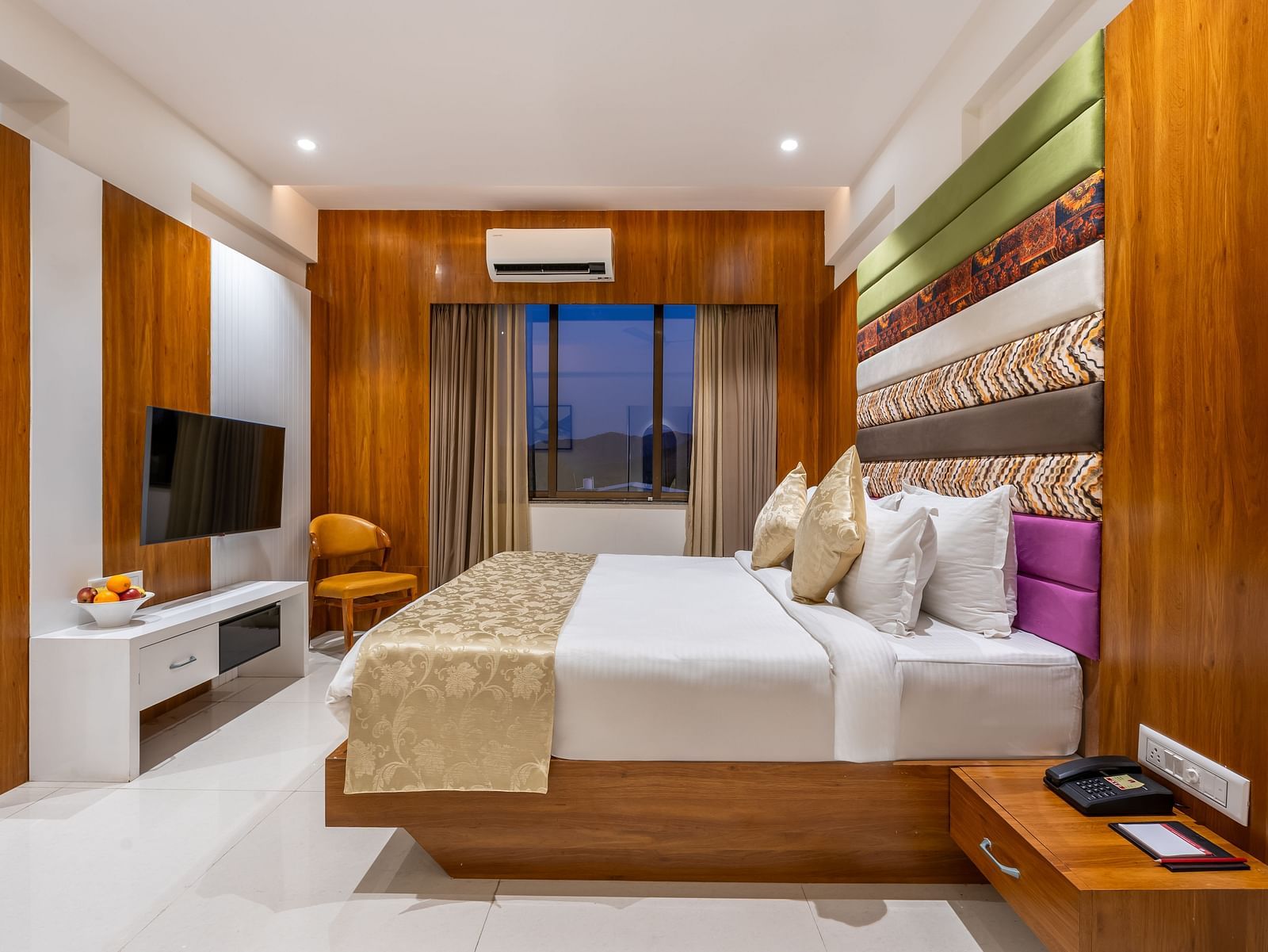 A hotel room showcases a large bed with decorative pillows and throws, a mounted TV, and a window with curtains - Lords Eco Inn Ambaji.