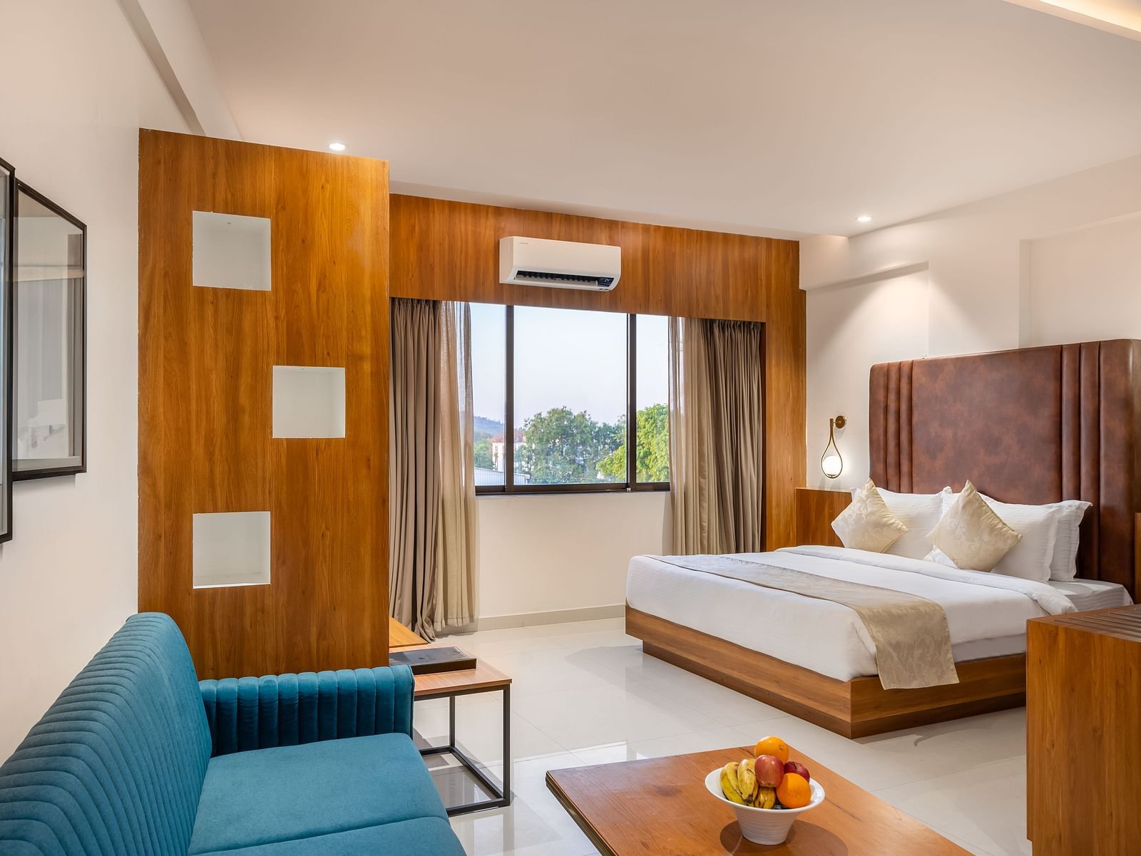 A well-lit hotel suite features a comfortable bed, a sofa, a wooden coffee table with fruit, and a window with curtains - Lords Eco Inn Ambaji.
