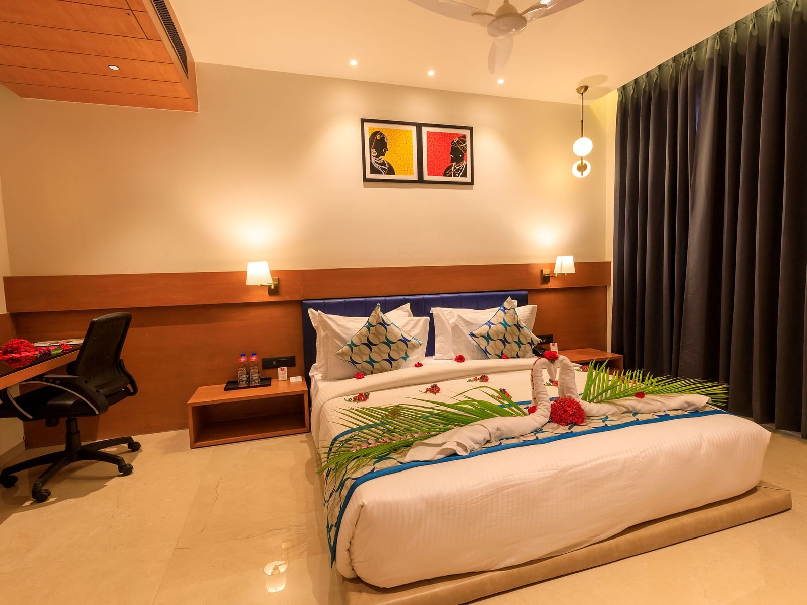 A warmly lit hotel room at Lords Eco Inn Apple Gate features a decorated double bed, bedside tables, a desk with a chair, and dark curtains.