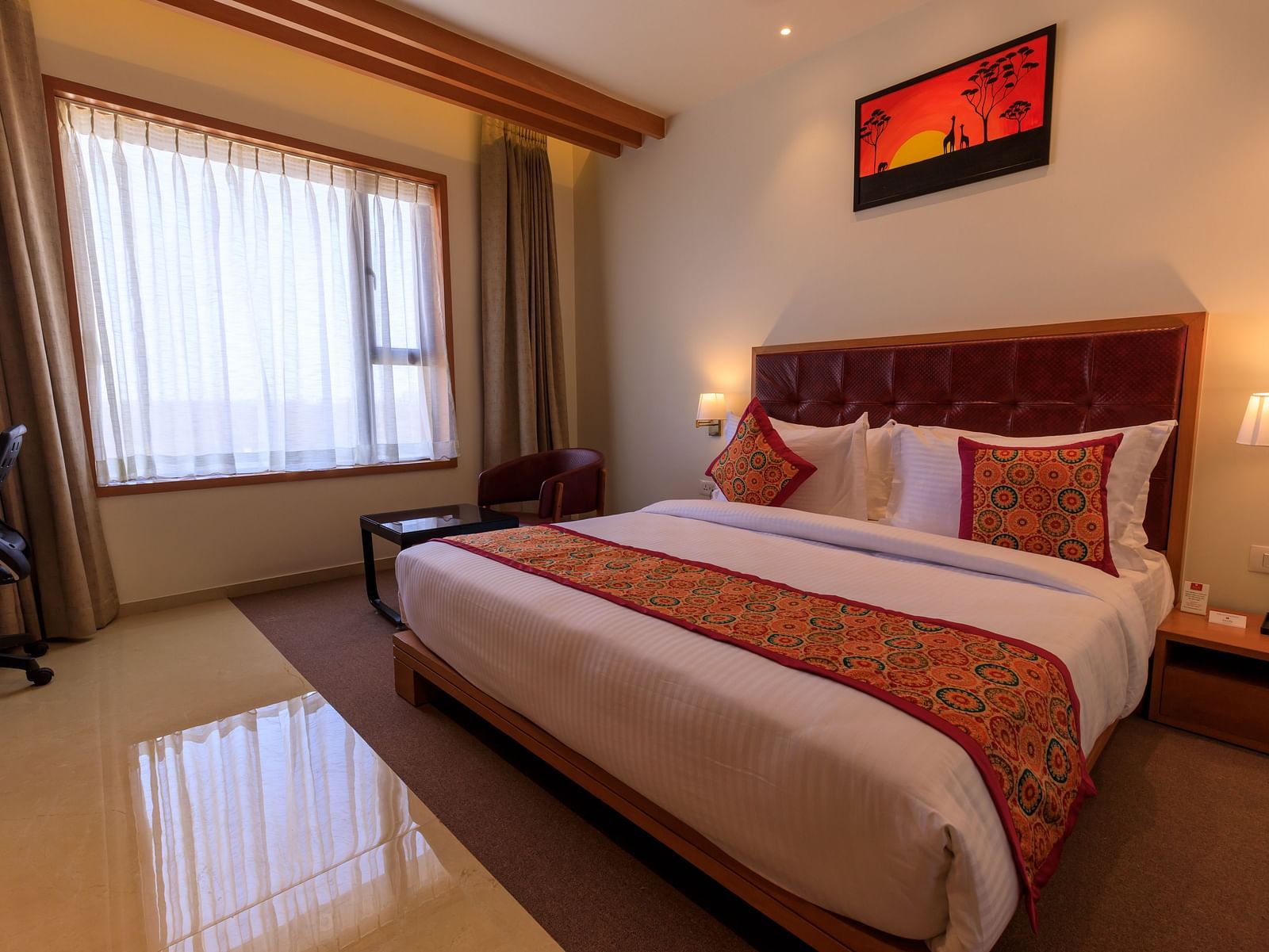 A well-lit hotel room at Lords Eco Inn Apple Gate features a double bed with patterned accents, a desk, a chair, and window curtains.