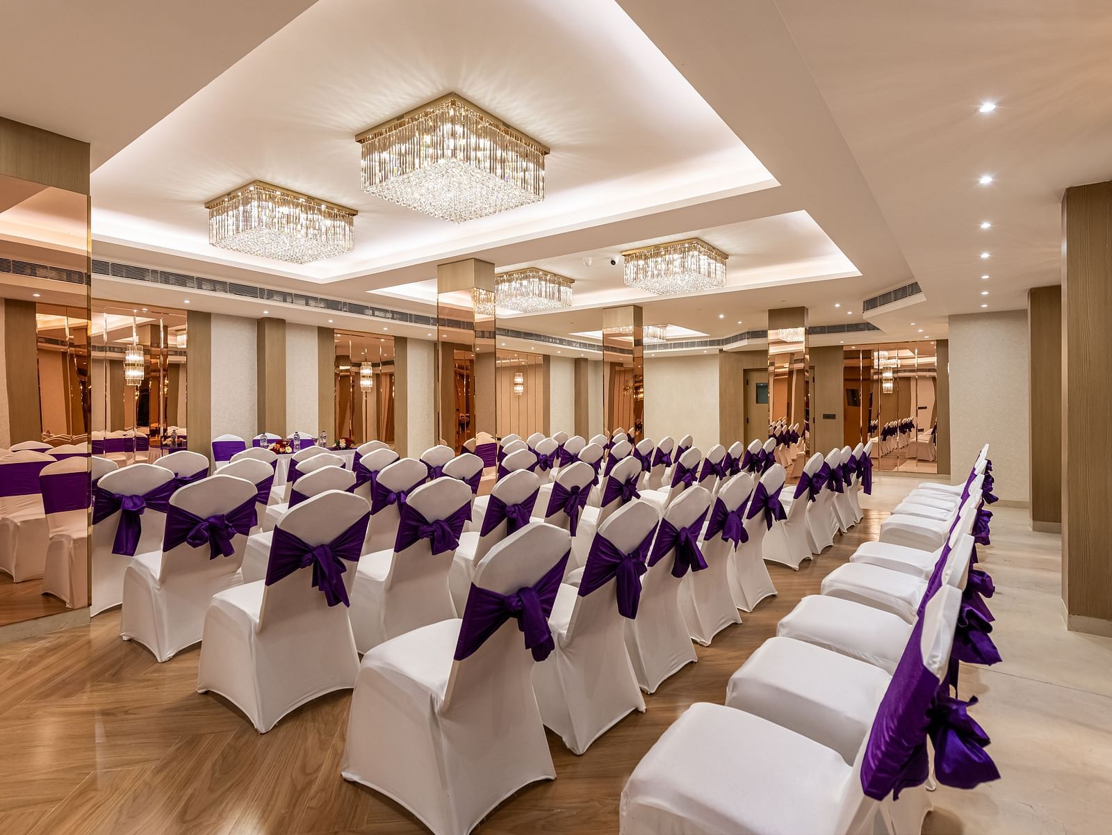 An event space with rows of white chairs decorated with purple fabric, ready for guests. It features a decorative ceiling with hanging lights and mirrored wall accents - Lords Eco Inn Dehradun.