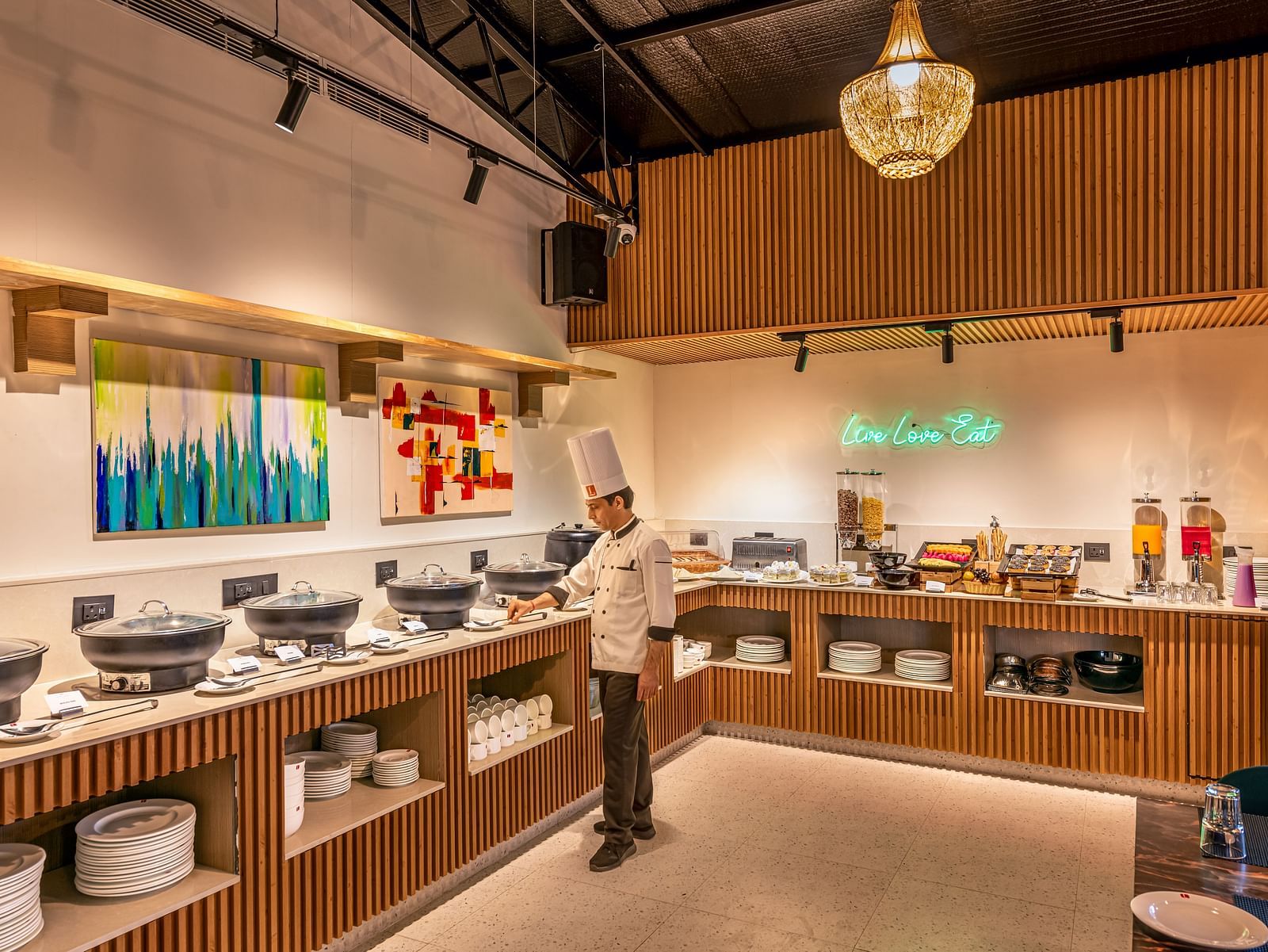 A buffet area with a chef standing near serving dishes, offering a variety of food and drinks. It features wooden paneling, a chandelier, and colourful artwork on the wall - Lords Eco Inn Dehradun.