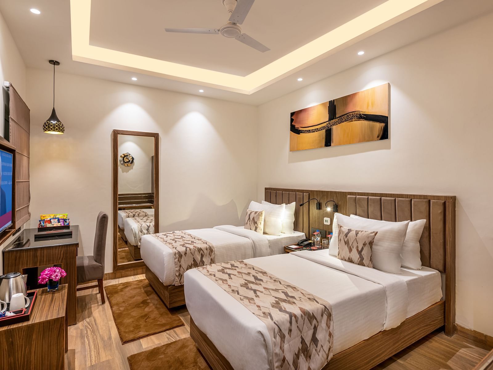 A hotel room with two twin beds, a flat-screen TV, and a work desk. It features a unique tray ceiling with lighting, a ceiling fan, and colourful abstract artwork - Lords Eco Inn Dehradun.
