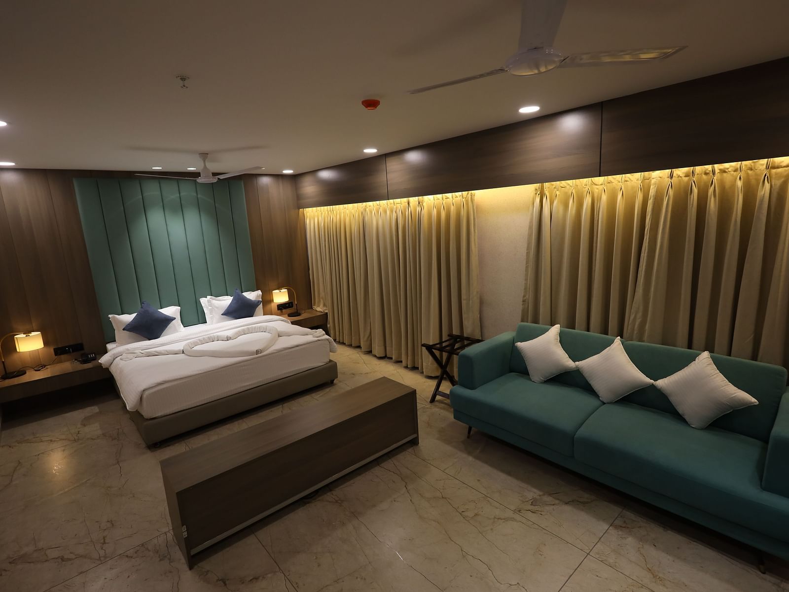 The image shows a spacious hotel room featuring a bed, a sofa with cushions, bedside tables with lamps, and full-length curtains - Lords Eco Inn Navsari.