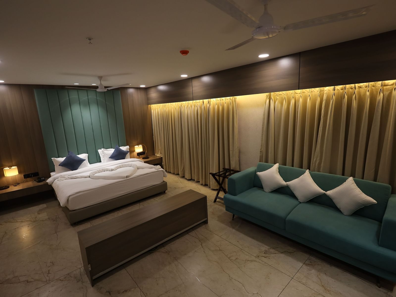 The image shows a comfortable hotel suite with a bed, a sofa with throw pillows, bedside lighting, and window coverings - Lords Eco Inn Navsari.