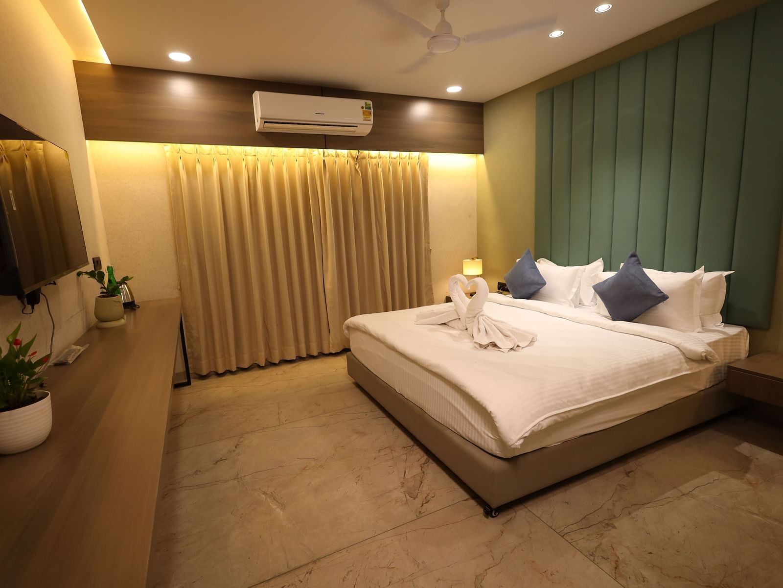 The image shows a well-lit bedroom with a bed, pillows, bedside lamps, curtains, and a dresser with plants - Lords Eco Inn Navsari.