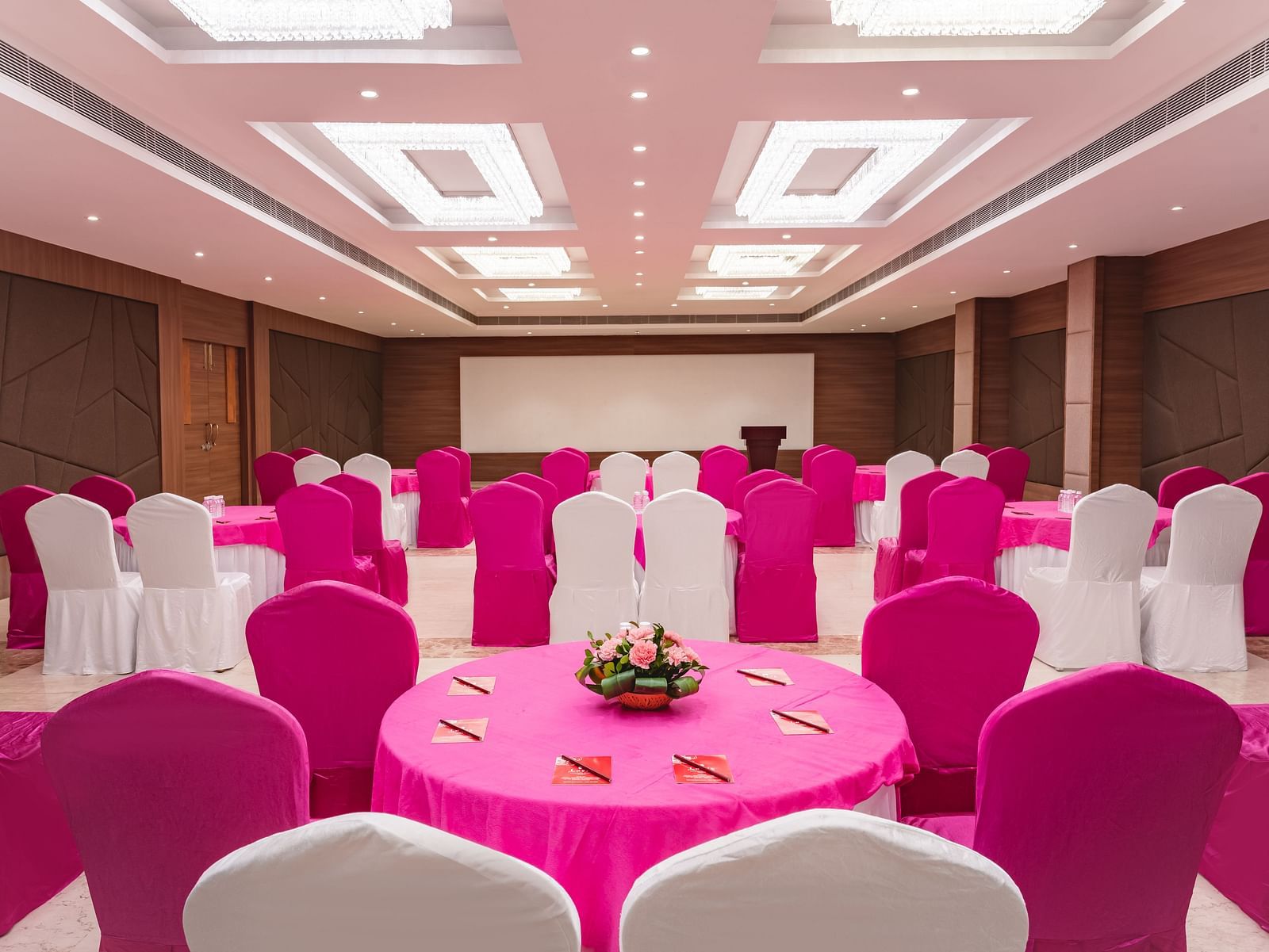 A banquet hall at Lords Inn Udaipur is set for an event with round tables draped in pink, surrounded by chairs in pink and white covers, facing a stage.