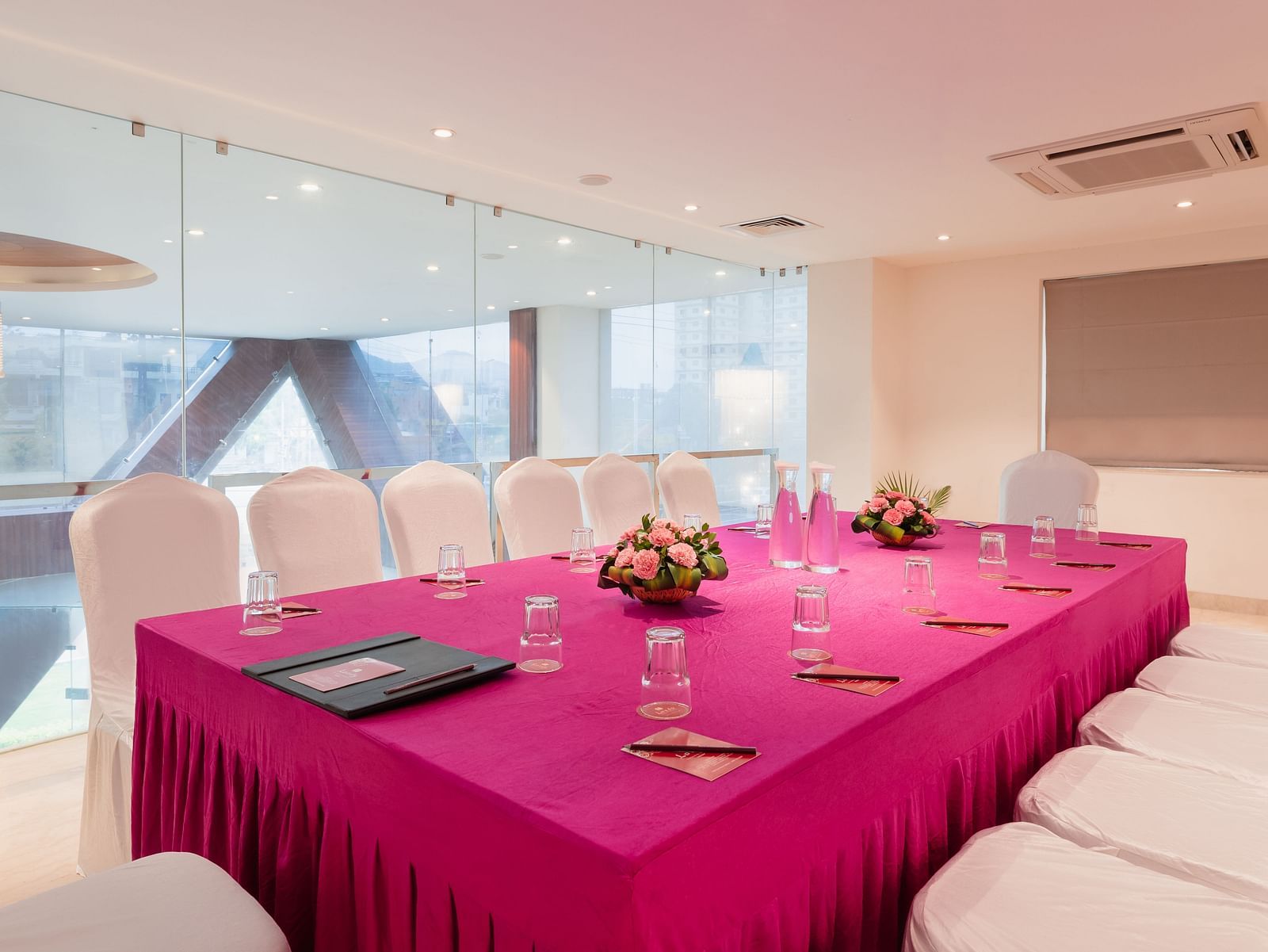 A meeting room at Lords Inn Udaipur is set with a long table covered in a vibrant cloth, surrounded by white chairs and adorned with floral centerpieces and water glasses.