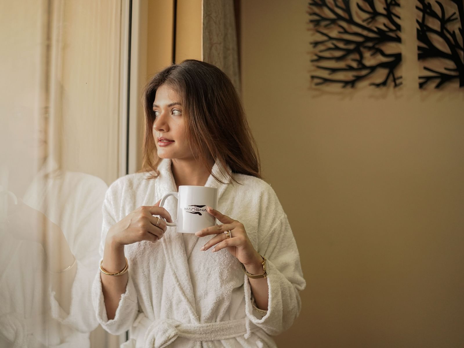 A woman in a white robe holding a cup near a window | Mansha Hotels