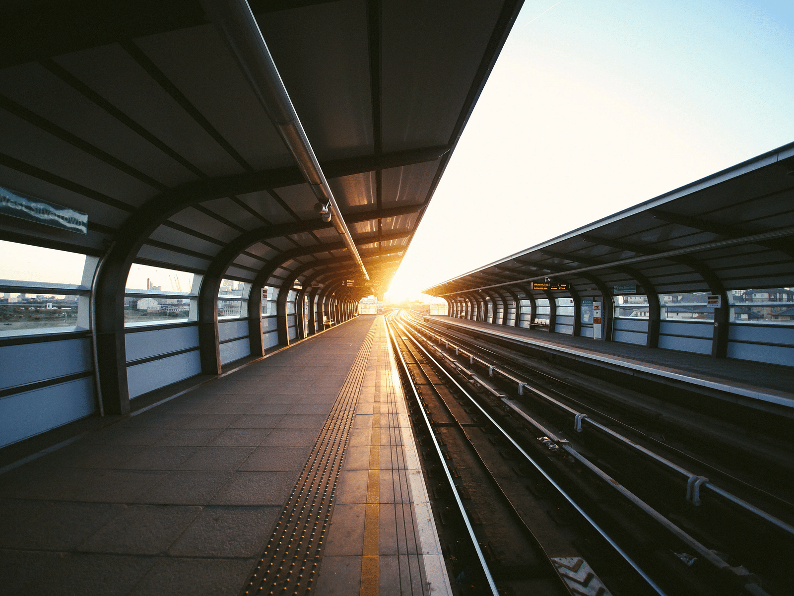 An empty train station with tracks in the centre and platform on the side and sun setting in the background