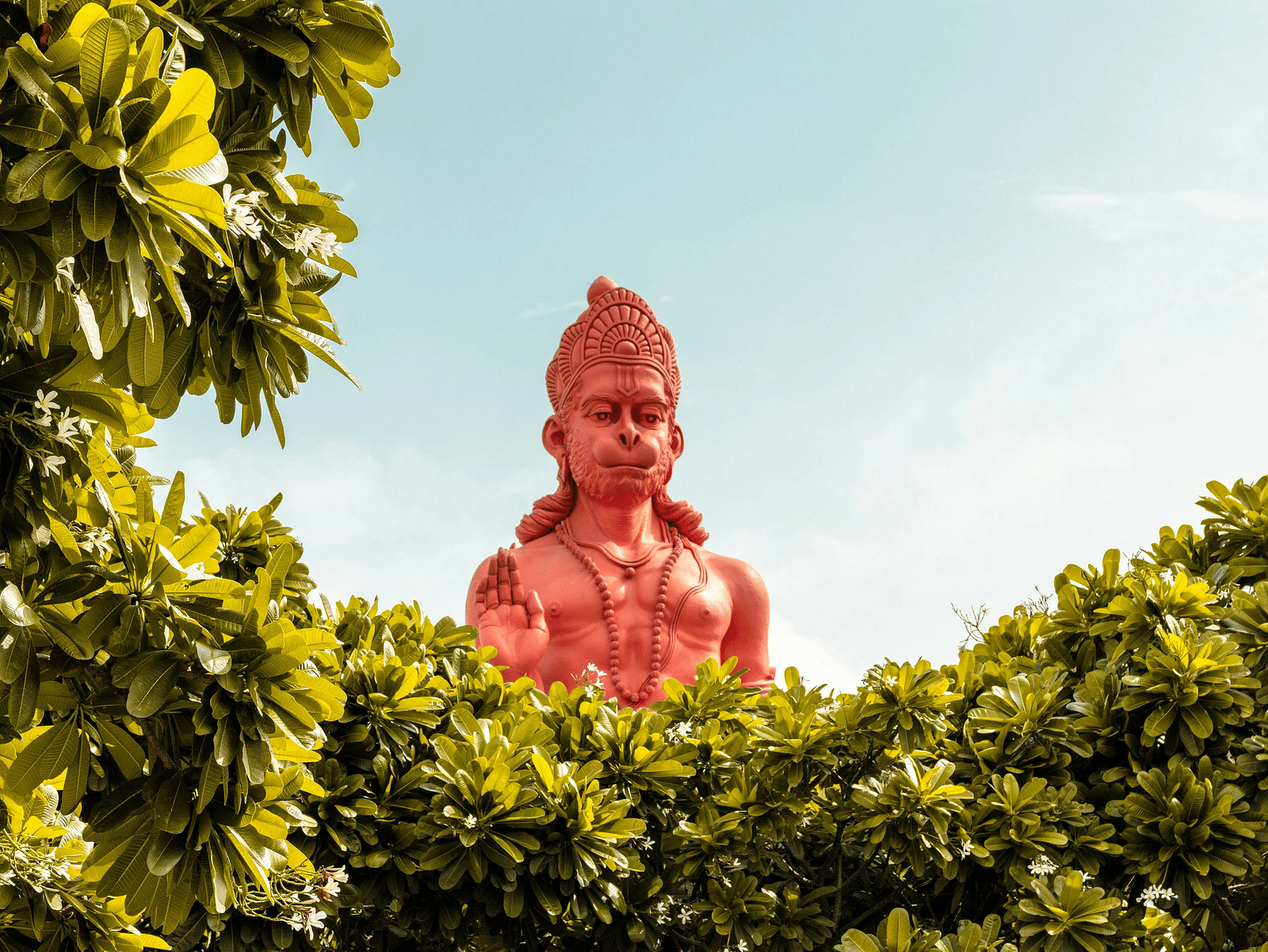 A towering red Hanuman statue emerging through lush greenery under a clear sky.