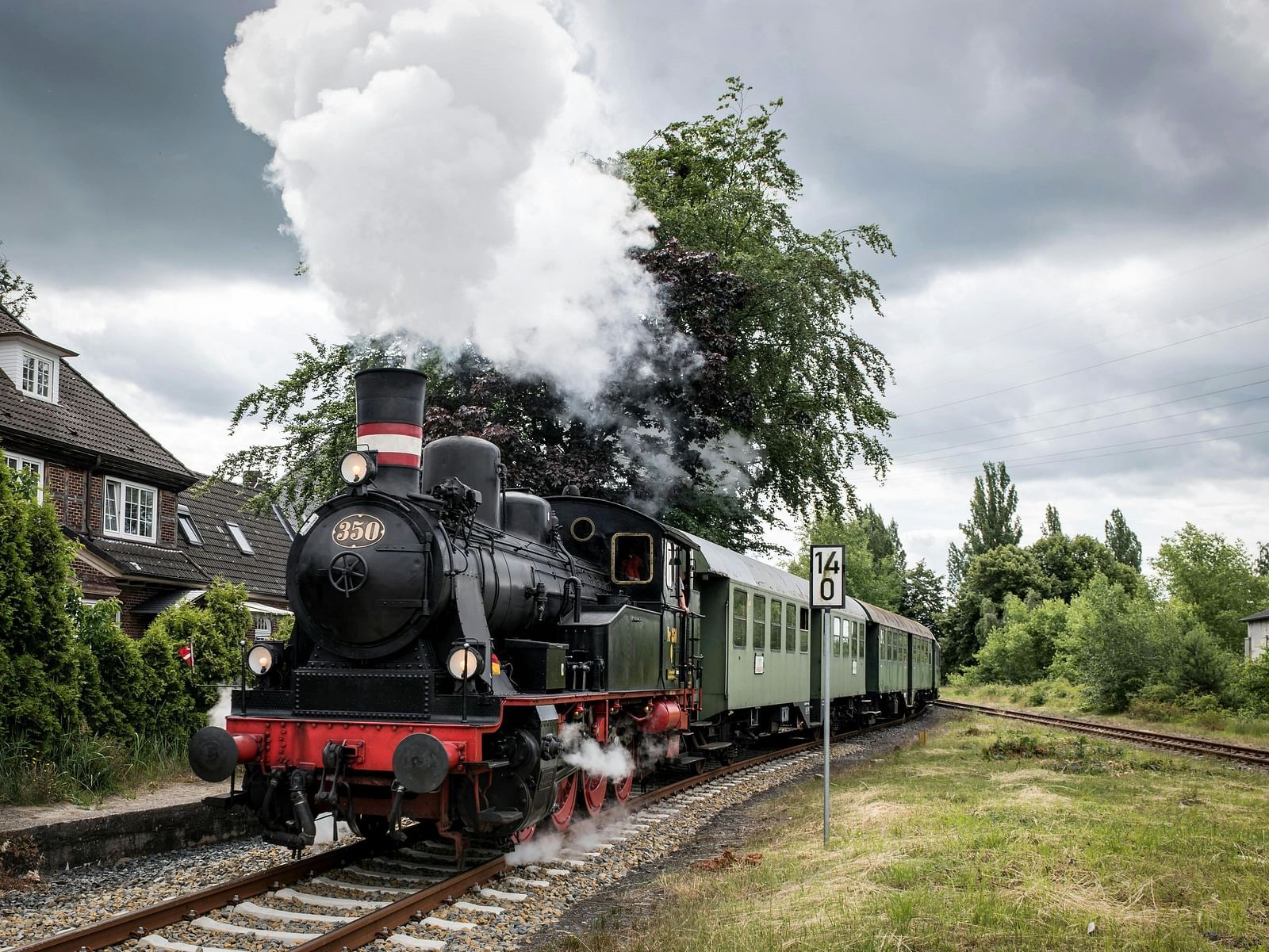 A vintage steam train passing by houses under a cloudy sky.