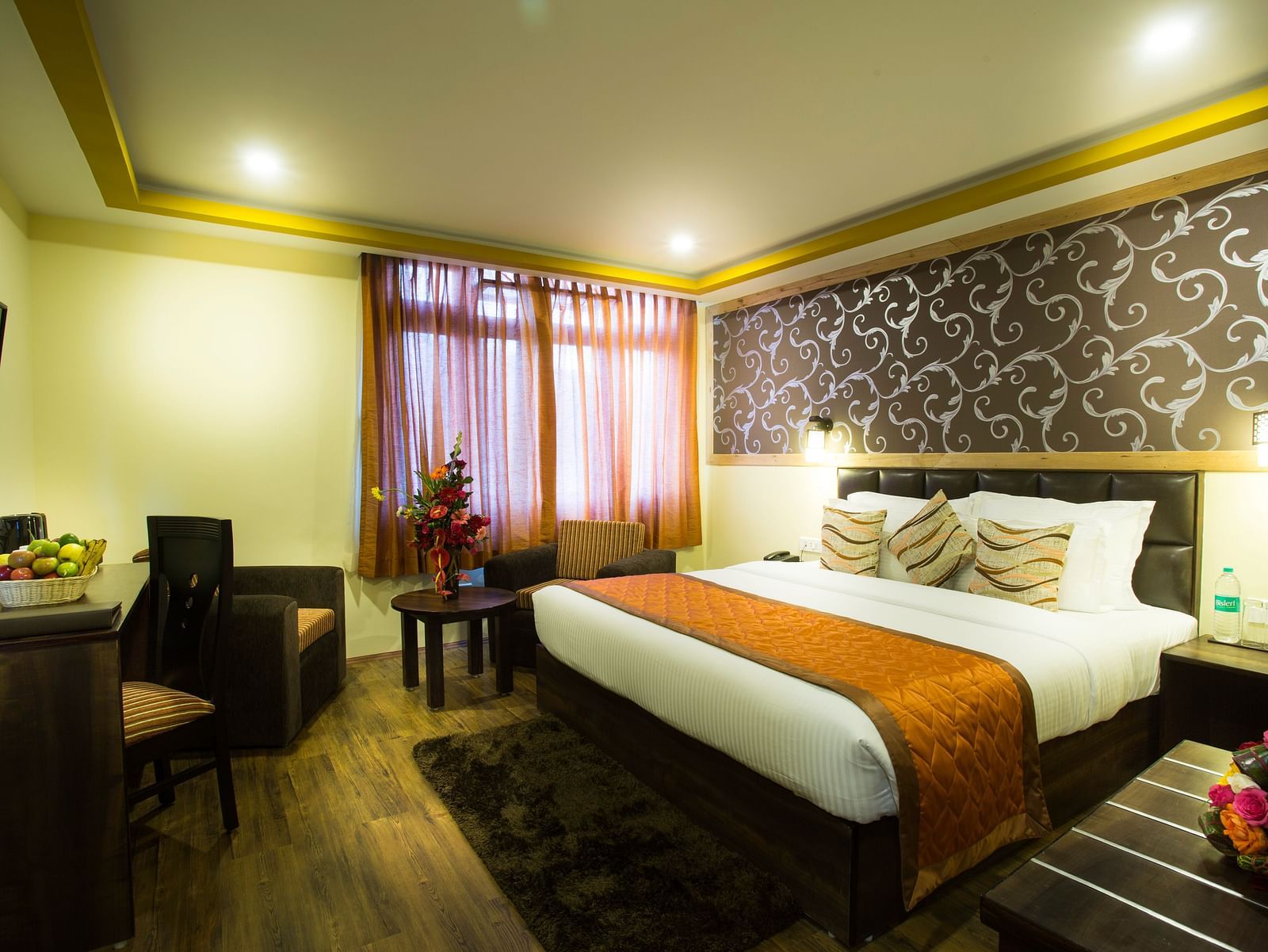 A  room at New Orchid Lords Inn Gangtok includes a comfortable bed with decorative pillows, a bedside table with water bottles, a functional desk with a chair, and a mounted TV for entertainment.