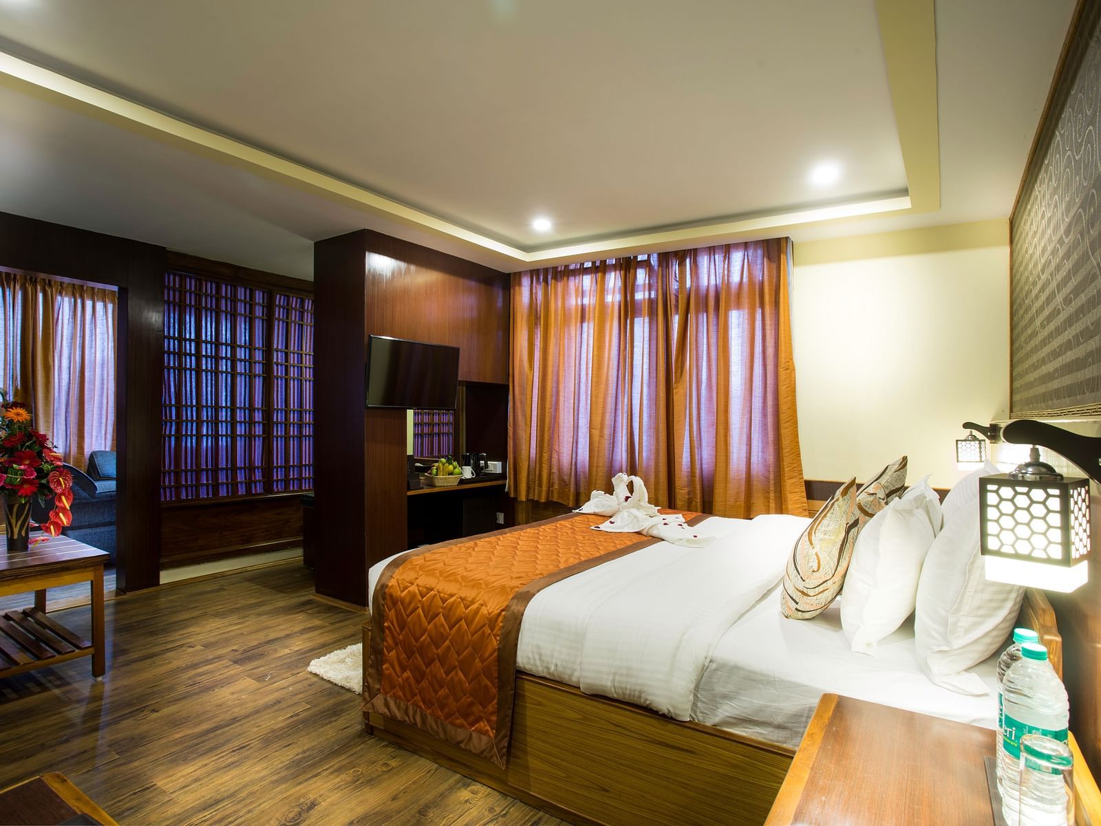 A room at New Orchid Lords Inn Gangtok contains a bed with pillows and folded towels, bedside tables with lamps and water, curtains, and a table with flowers.