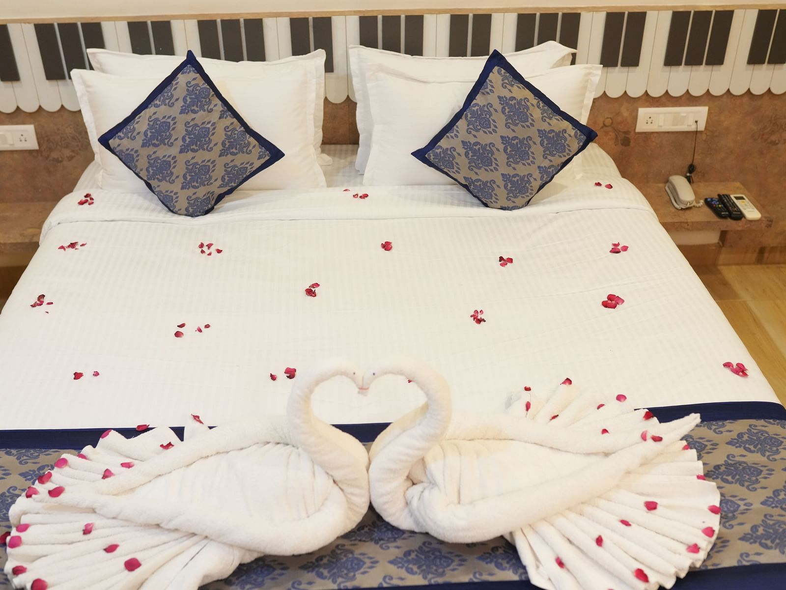 Romantic hotel room setup with white bedding decorated with scattered red rose petals, blue patterned decorative pillows, and swan-shaped towel art at SeaFORT Beach Resort