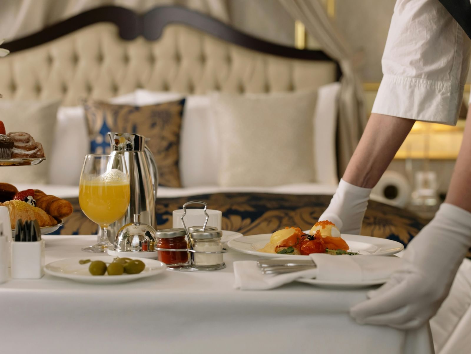 A hotel staff member meticulously serving a luxurious in-room breakfast spread on a bed, featuring various dishes and beverages.