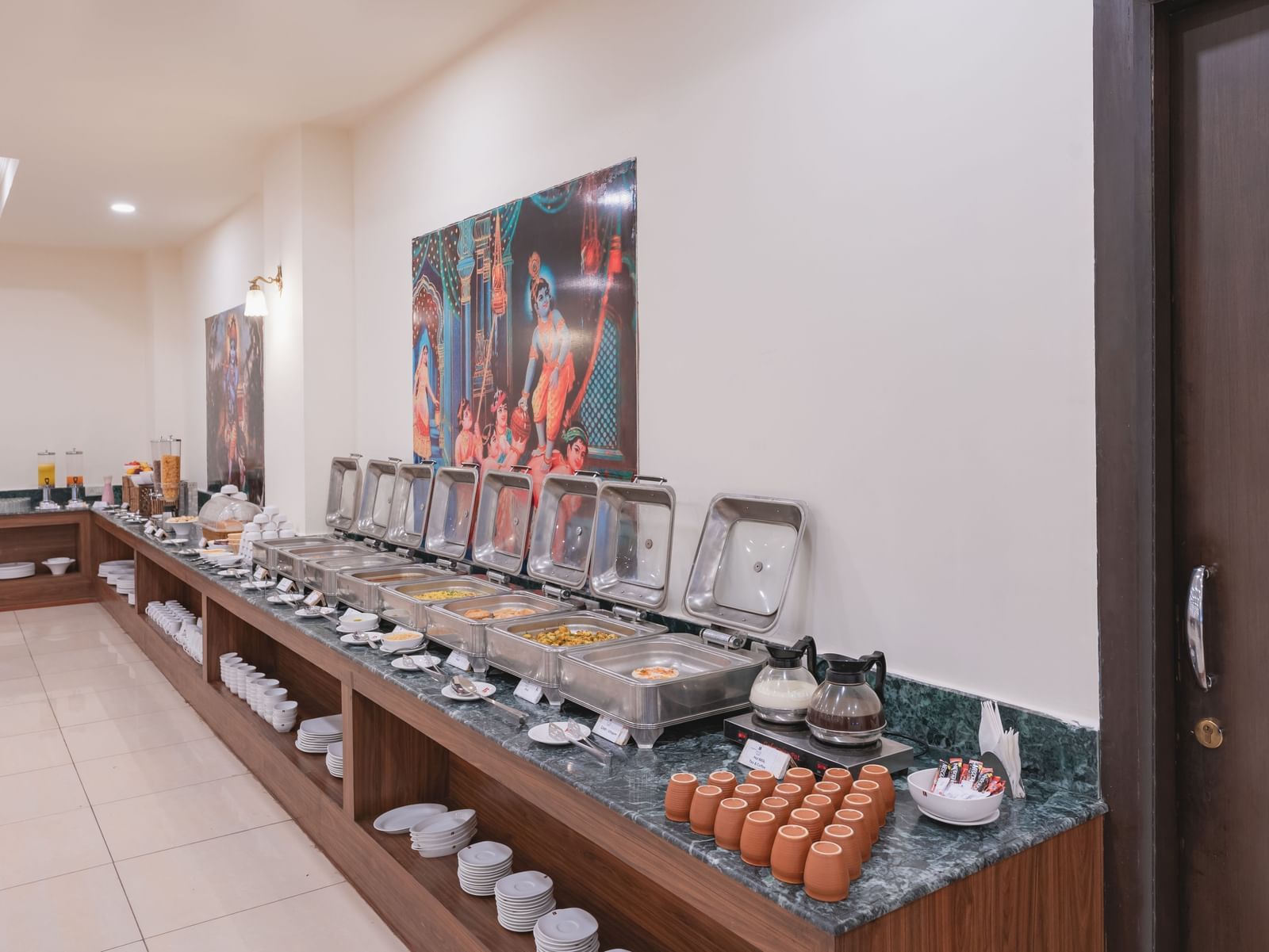 Buffet setup with covered serving dishes, beverages, and utensils arranged at the Manorath Restaurant at Shri Vallabh Vilas Lords Plaza Nathdwara, along a counter in a bright dining area.