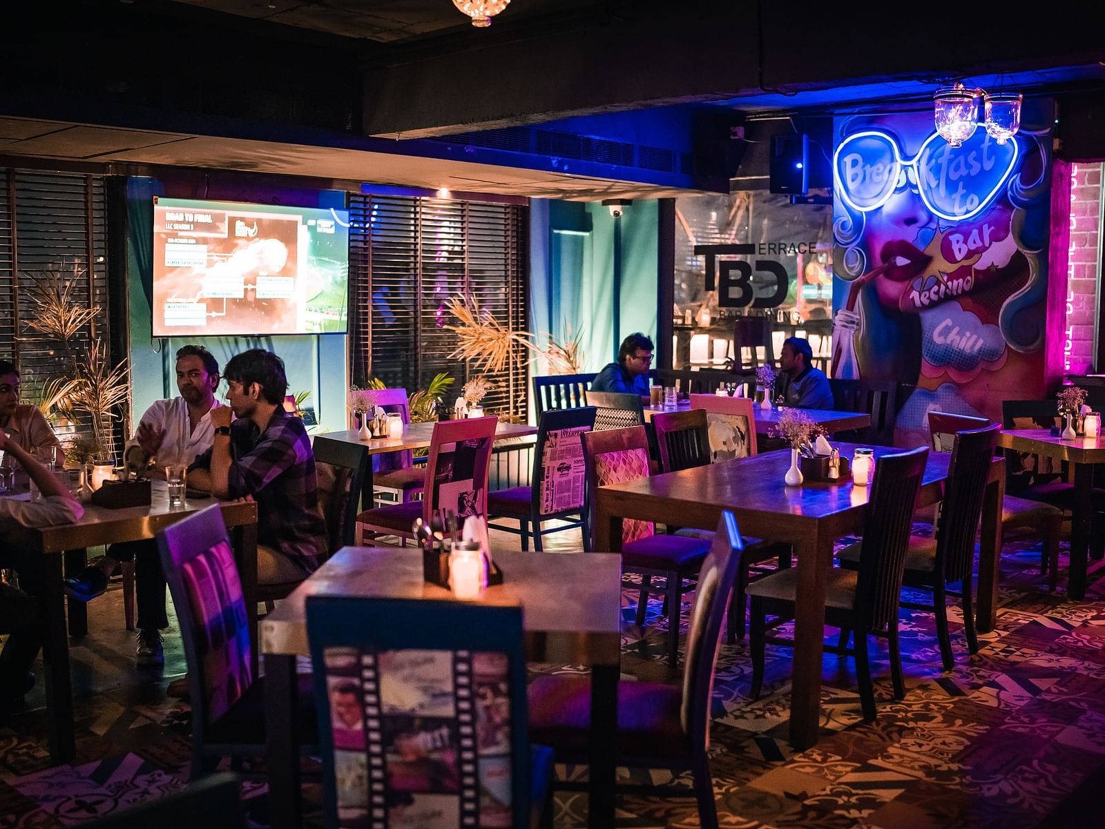 A lively, dimly lit bar with wooden tables and chairs, occupied by groups of people enjoying drinks and conversation. The walls are decorated with neon signs and eclectic artwork, creating a vibrant and energetic atmosphere.