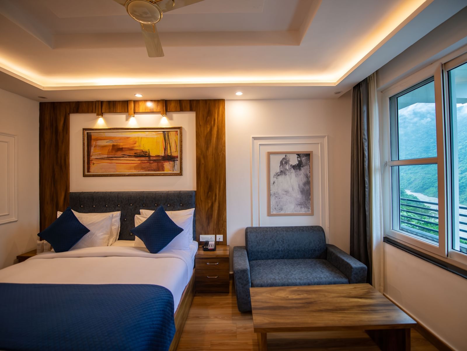 A Deluxe Room at The Tattva Devaprayag with a bed featuring blue blankets and two pillows, two framed paintings on the wall and a sofa.