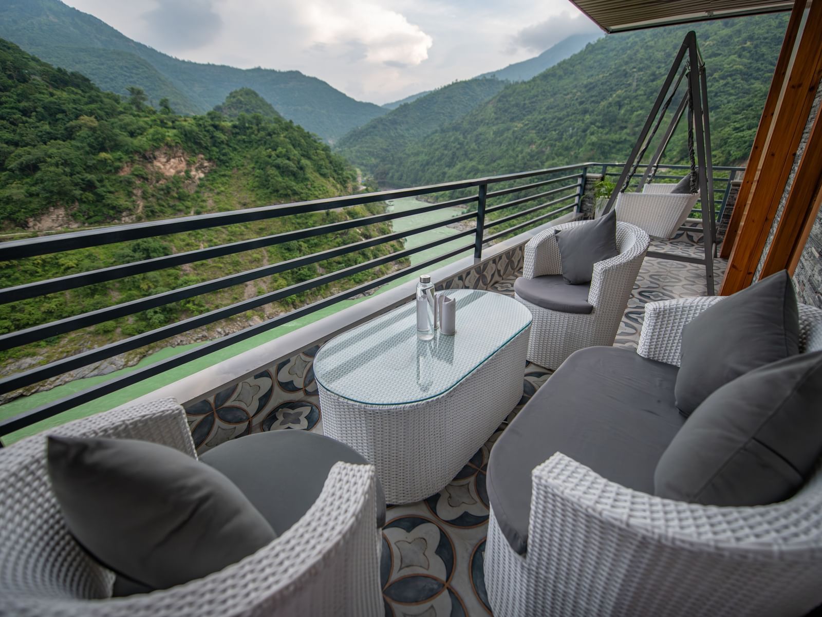 A wide view of mountains and river flowing, a seen from the balcony of The Tattva Devaprayag featuring two grey chairs, a table and a swing.