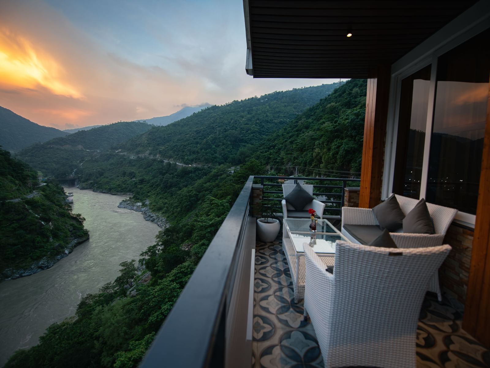  A wide view of the mountains and a river as seen from the balcony of The Tattva Devaprayag featuring a white couch and a table during sunset.