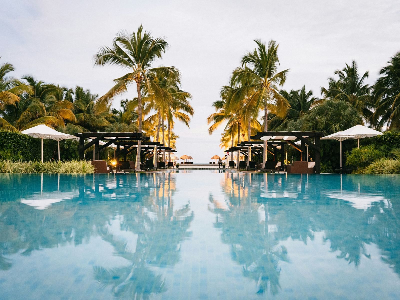 Expansive resort swimming pool reflecting the sky and surrounding palm trees and cabanas.