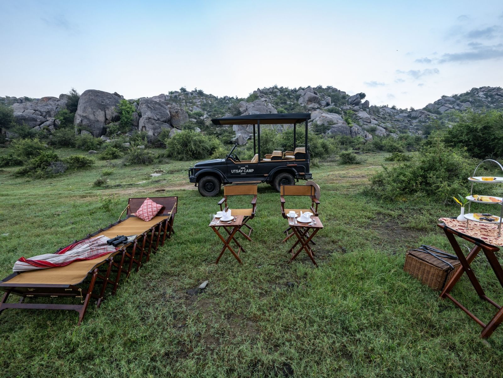 An image of an outdoor seating arrangement with a jeep parked nearby amidst greenery and the backdrop of green mountains - Utsav Camp Sariska