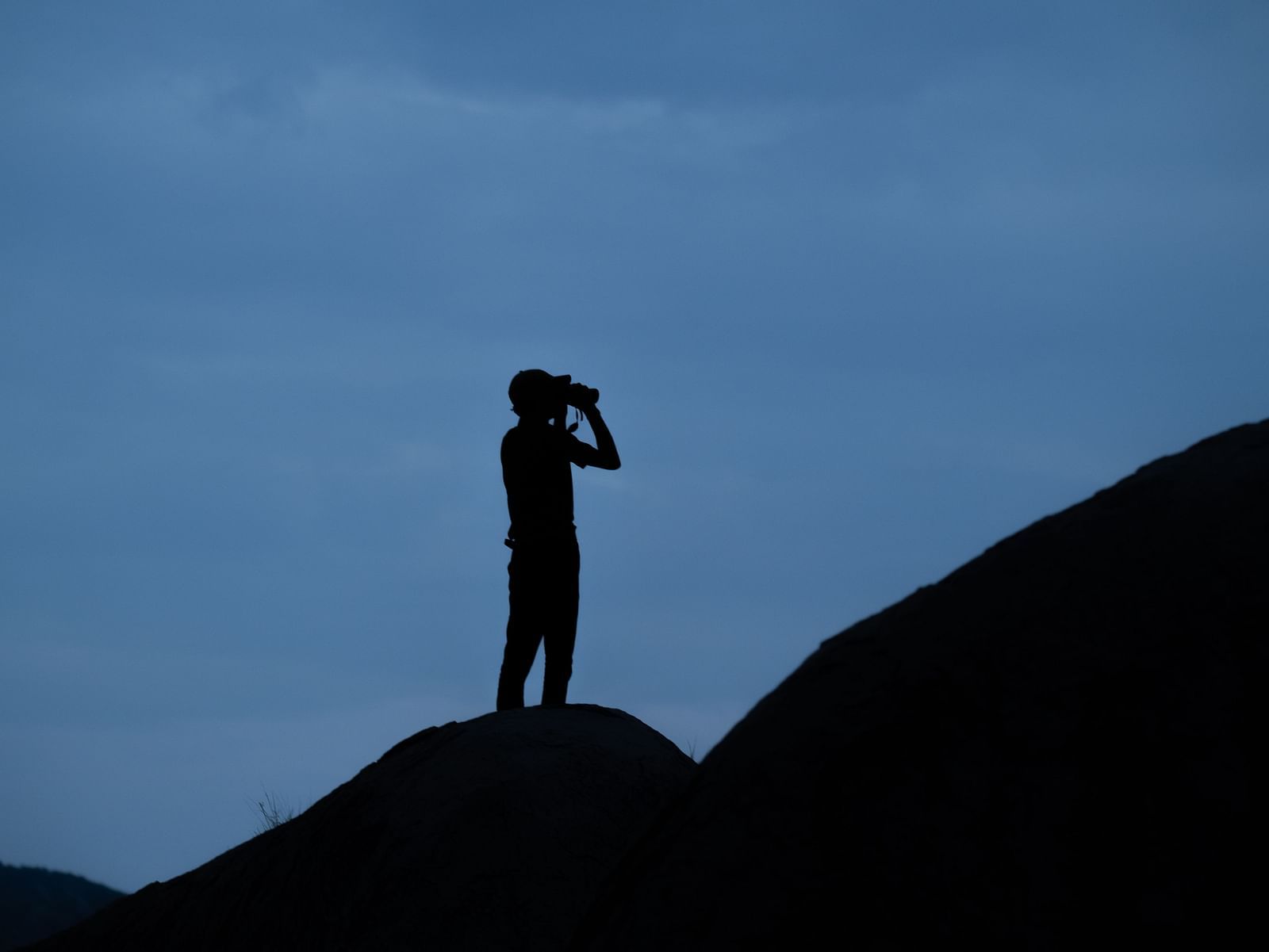 Person against a twilight sky, standing on a rocky outcrop, holding binoculars.