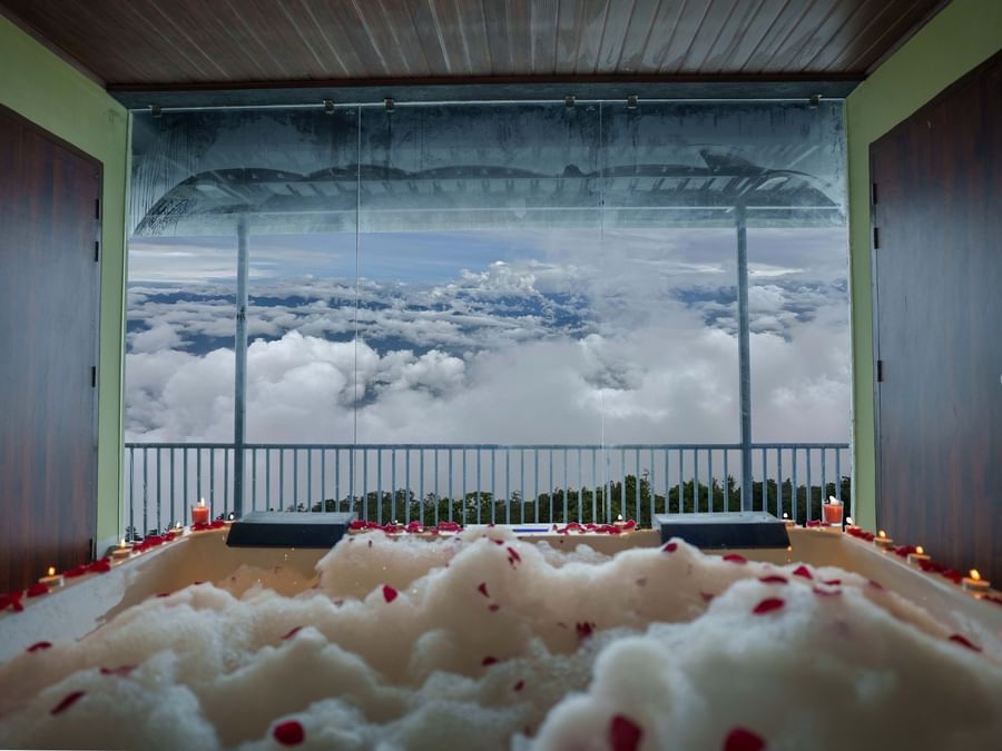 A jacuzzi with rose petals scattered, leading to a balcony overlooking a cloudy sky and distant hills, suggesting a romantic setup at Summit Enigma Resort and Spa.