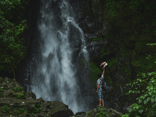 A person with a rucksack stands before a tall, misty waterfall cascading down a rocky cliff in a lush green forest.
