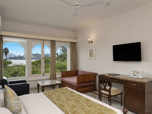 A guest room at Ram Pratap Palace with a bed, pillows, two chairs, a coffee table, a desk, a television, a lamp, and a window with arched frames overlooking the lake.