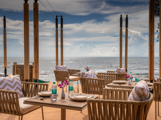 A well arranged tables and chairs at In-House Restaurant with sea waves crashing on the beach in the background