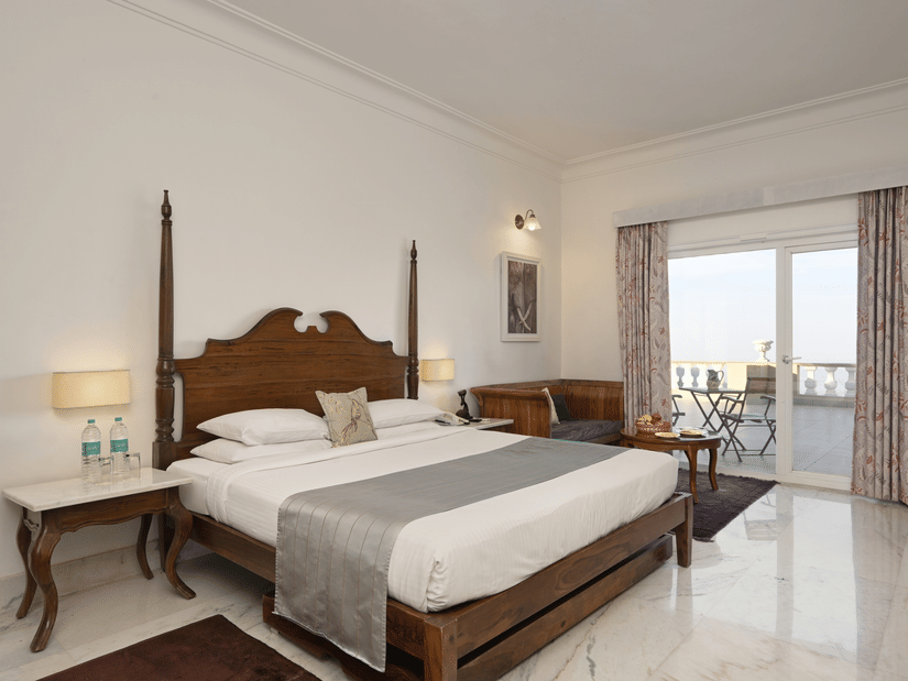 Deluxe Room with Balcony at Fateh Safari Resort, Kumbhalgarh featuring a double size bed, bedside table with water bottles, a seating area, and a balcony attached which provides an outdoor vide.