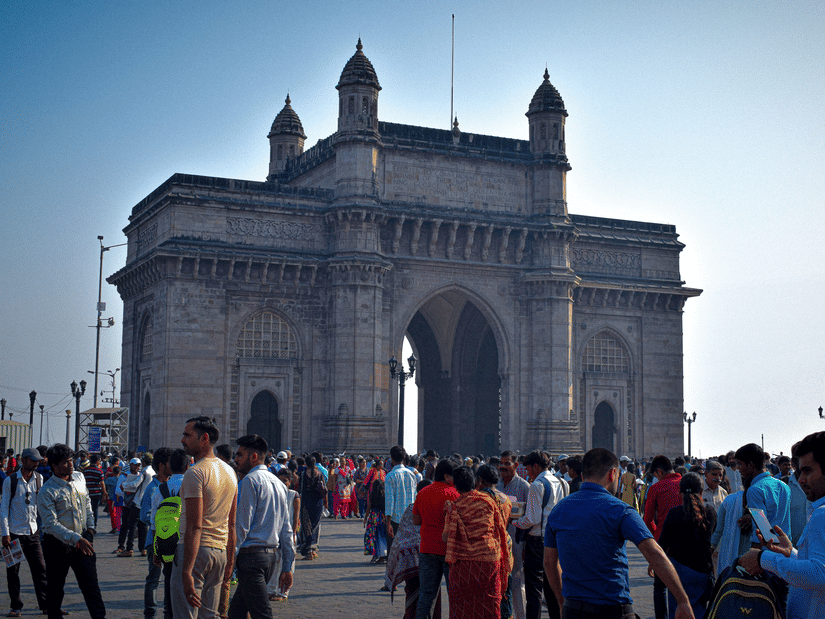 Full view of the Gateway Of India historic monument on a bright sunny afternoon with huge number of pedestrians in the foreground.