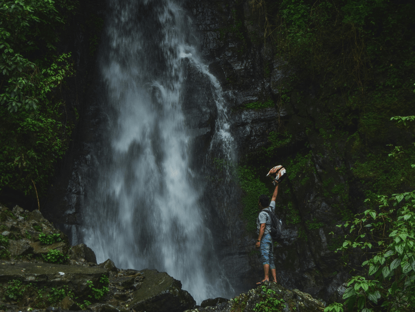 A person with a rucksack stands before a tall, misty waterfall cascading down a rocky cliff in a lush green forest.