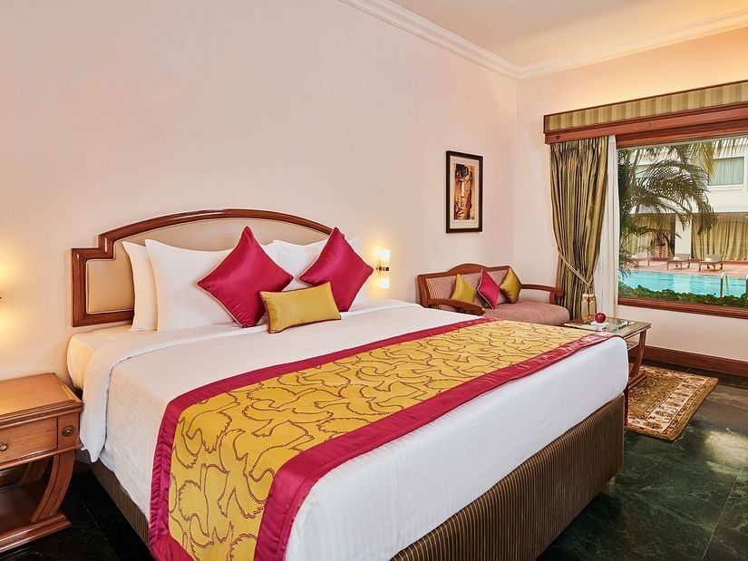 Luxurious hotel room at The Ummed Ahmedabad featuring elegant bed with colorful pillows, warm lighting, and modern furnishings in beige tones.