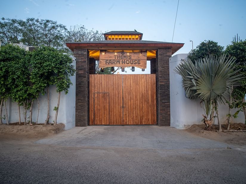 Grand wooden entrance gate of Themis Farm House, flanked by lush greenery and palm trees, set against a serene, blue-sky backdrop.