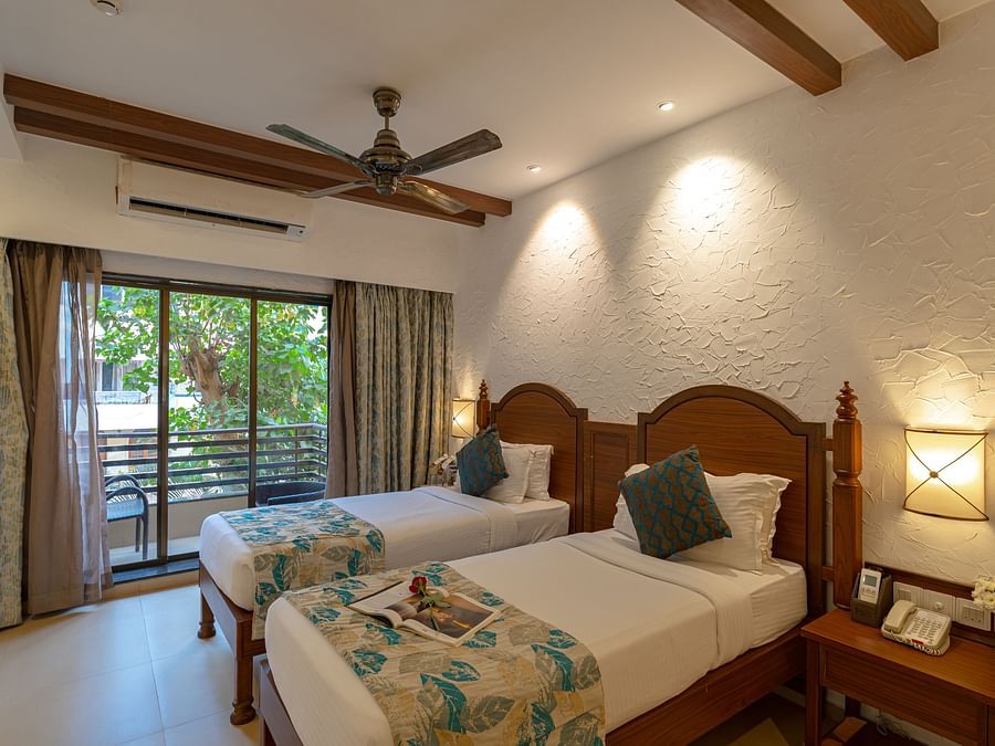 An image of a hotel room with twin beds dressed in white sheets, a cosy throw, wall-mounted lamps on each side, and an attached balcony.