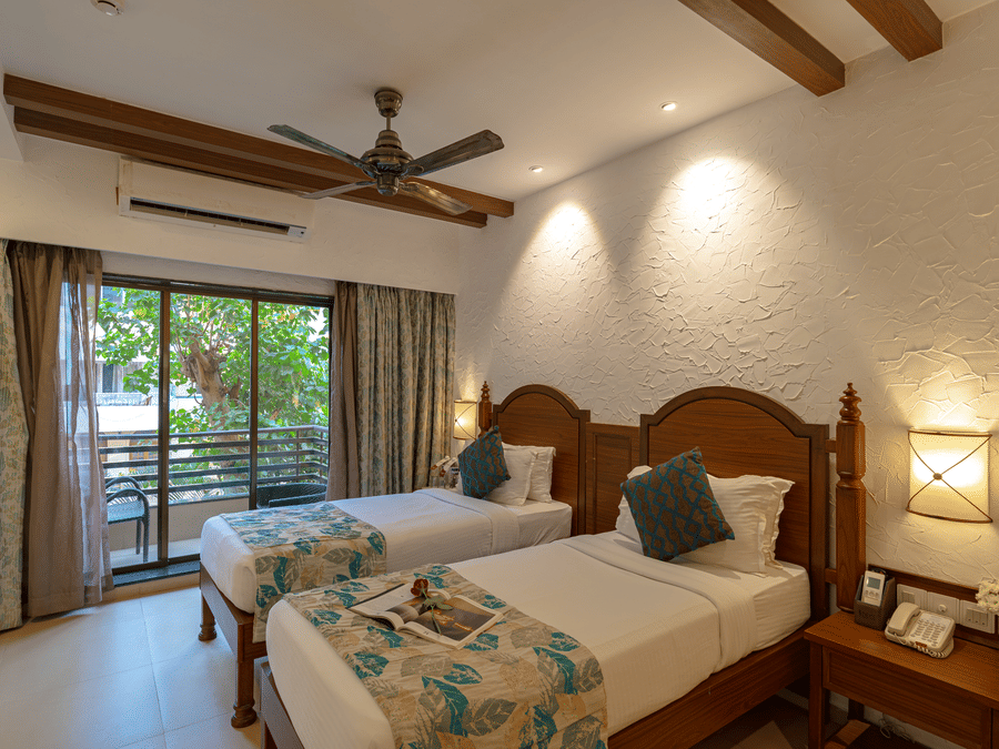 An image of a hotel room with twin beds dressed in white sheets, a cosy throw, wall-mounted lamps on each side, and an attached balcony.