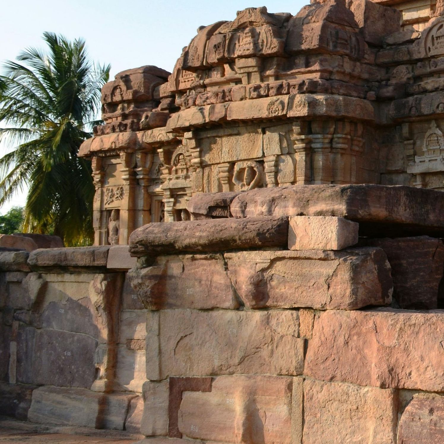 An ancient stone temple complex with intricate carvings, set against a clear sky and surrounded by palm trees.