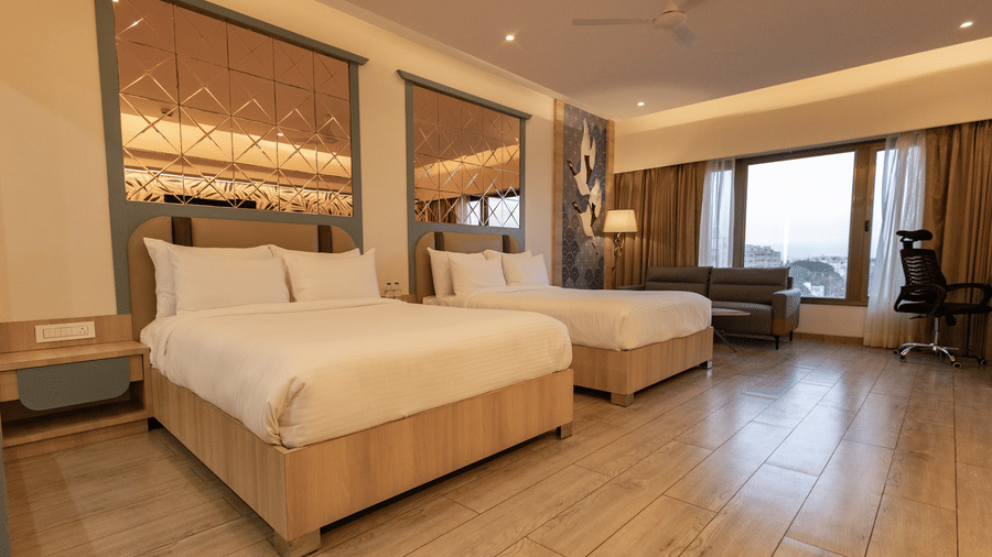 A well-lit hotel room featuring twin beds, a sofa near a window, wooden flooring, and a patterned headboard or wall panel | Pride Elite Chhatrapati Sambhajinagar