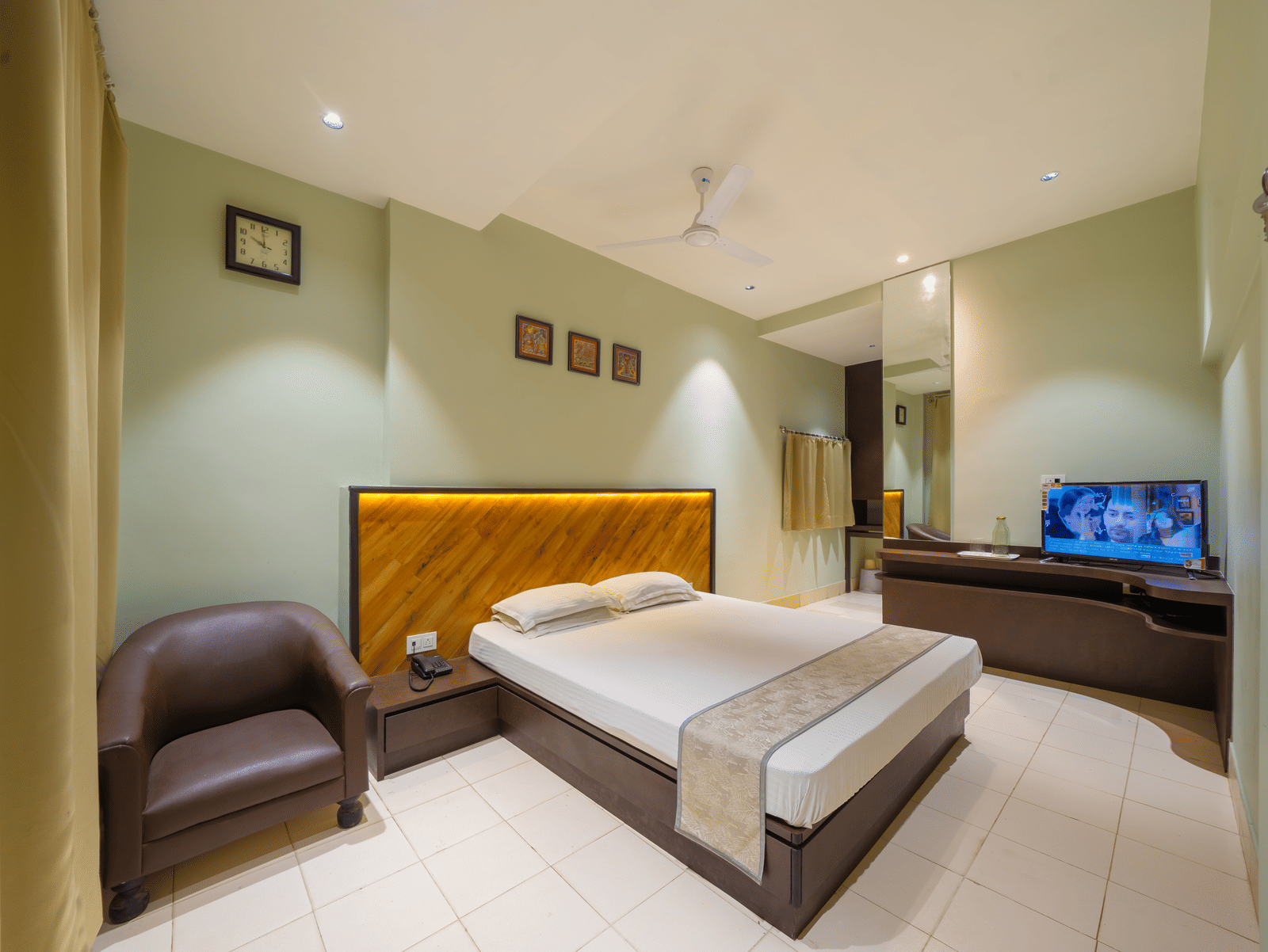 A spacious hotel bedroom featuring a double bed with a headboard, light green walls, a TV, and a leather armchair - Hotel Dolphin Puri