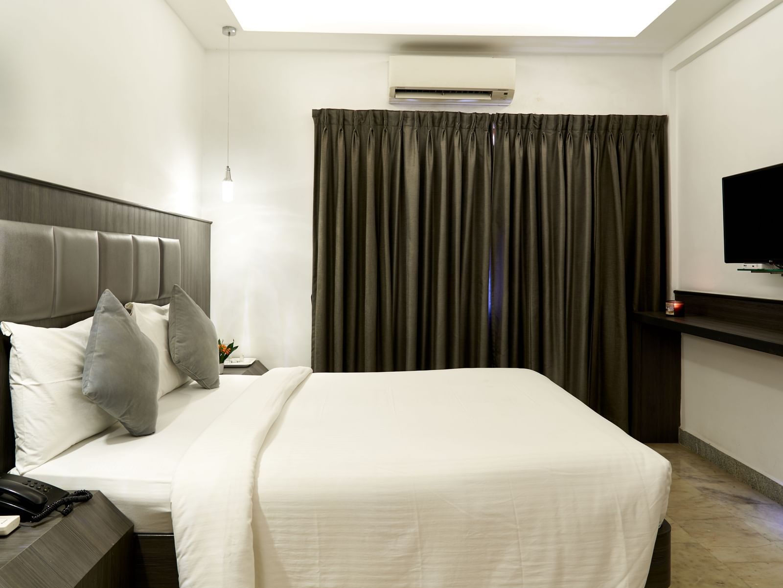 A modern hotel room at Hotel Mamallaa Heritage, featuring a bed with a dark upholstered headboard, white linens, and a flat-screen TV.
