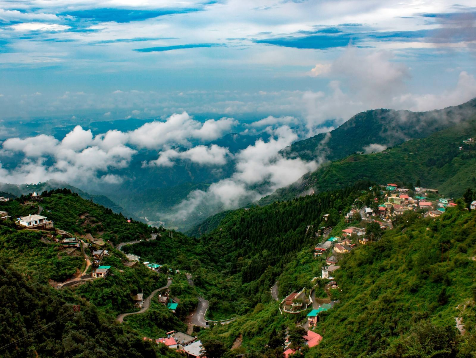 A wide-angle shot of a beautiful landscape of featuring hills covered with lush greenery along with clouds floating around in the vast blue sky.