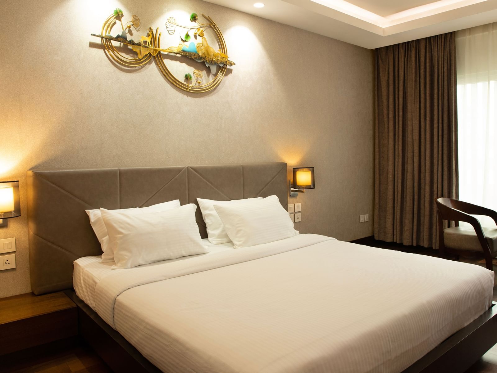 A bedroom with a large bed, bedside lamps, curtains drawn to the side, and natural light coming through the window at The Hamuse Hotel, Kodaikanal.