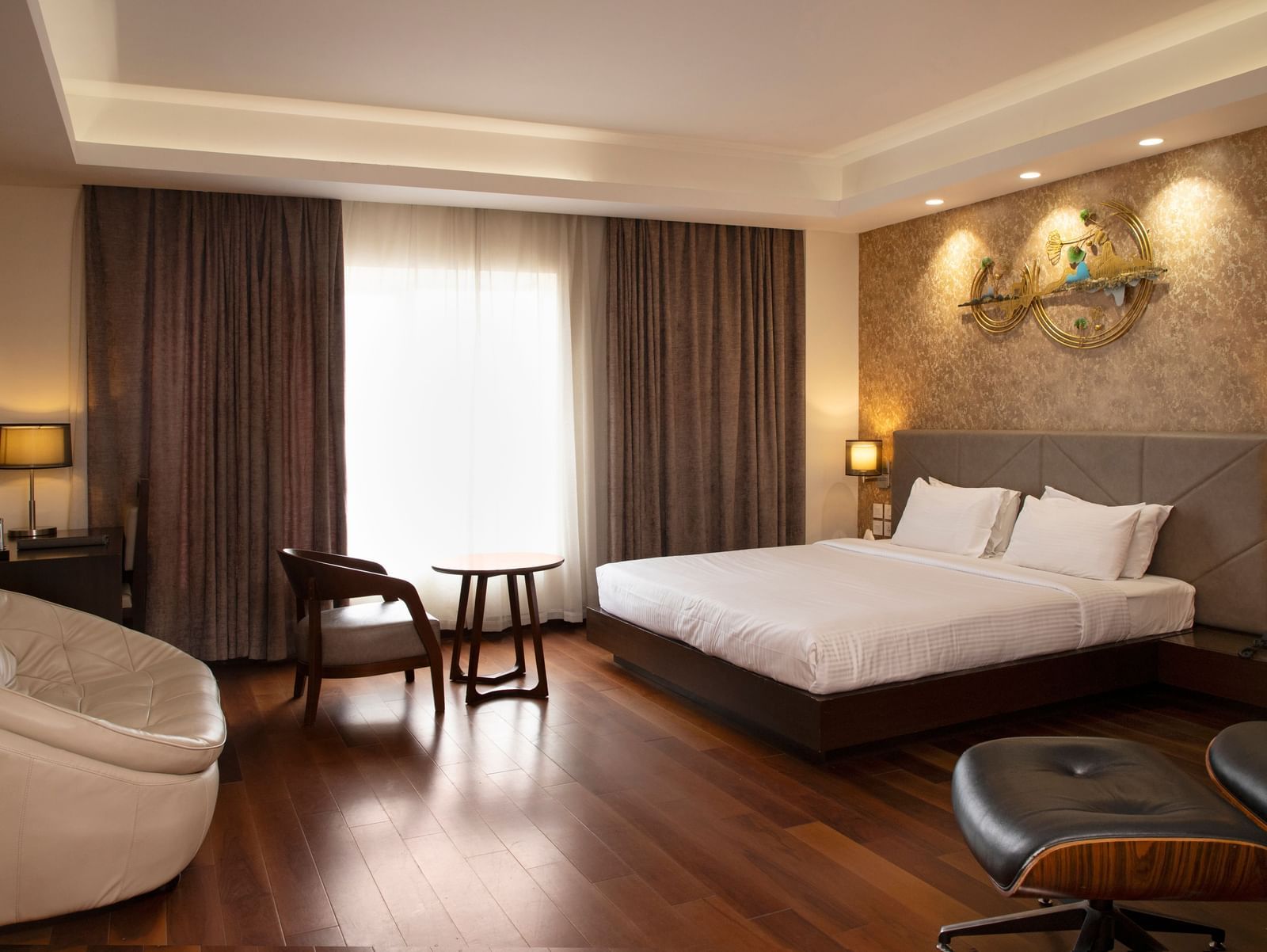 A spacious room featuring a large bed, a cosy seating area, elegant curtains, soft ambient lighting, and expansive windows with curtains drawn to the side at The Hamuse Hotel, Kodaikanal.
