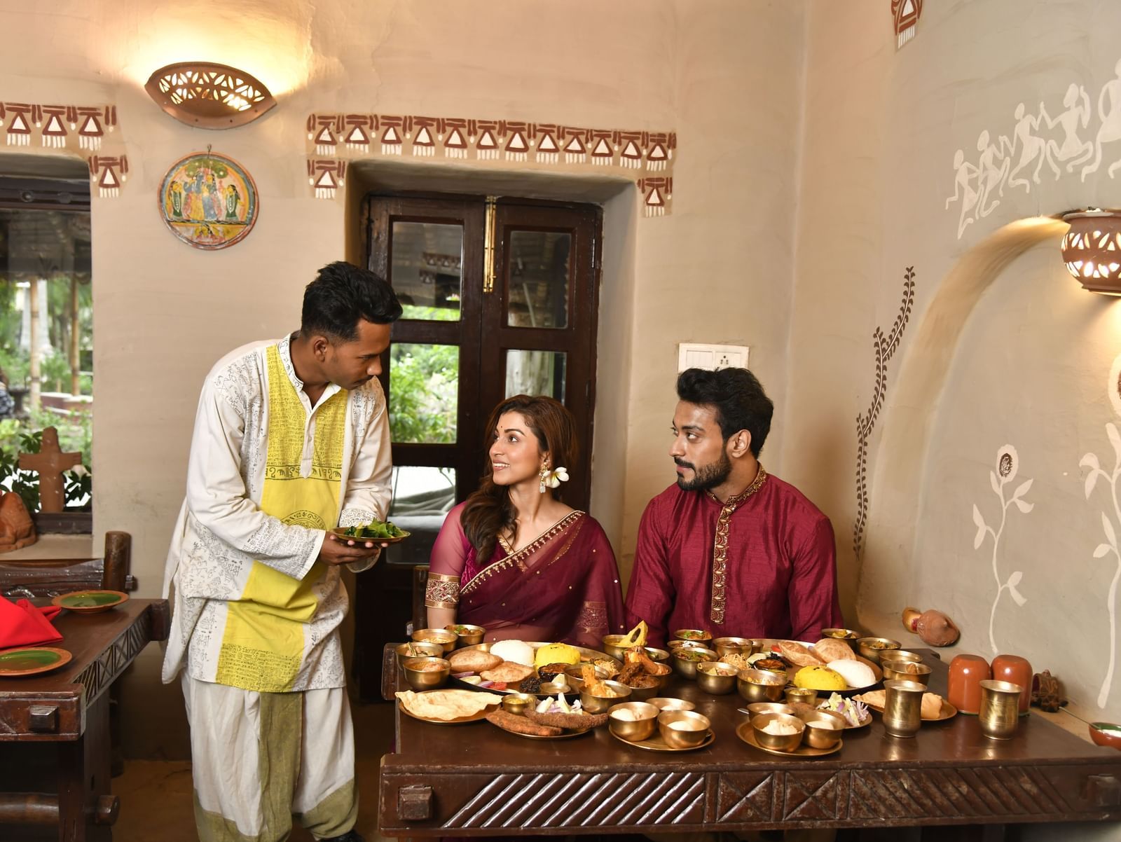 A server in traditional attire presenting food to two seated individuals at a low table | Bhoomi Restaurant